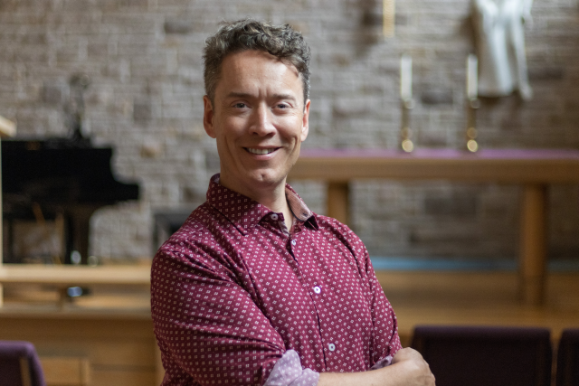 Greg Simpson smiling in the chapel, with his arms folded and a piano behind him